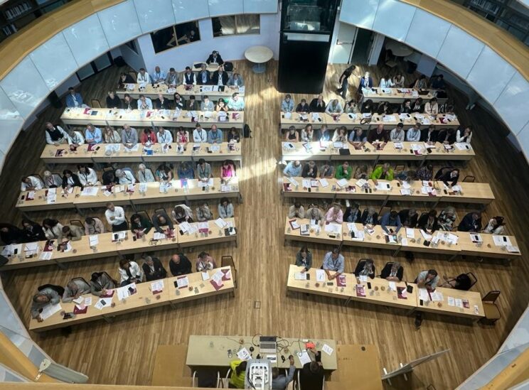 Vista cenital de asistentes al Encuentro de Consejos de Dirección de Hermanas Hospitalarias, sentados en mesas largas en una sala