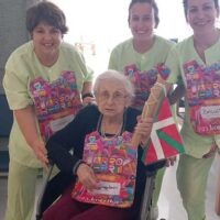 Tres mujeres auxiliares de enfermería acompañan a una mujer mayor en silla de ruedas que sostiene el testigo de la Korrika